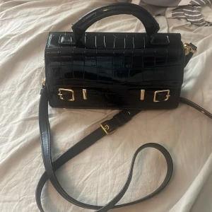 Svart axelväska med krokopräglat mönster - Snygg svart axelväska i glansigt krokopräglat material med guldfärgade spännen och detaljer. Väskan har både handtag och justerbar axelrem, samt magnetlås för smidig öppning. Perfekt för dig som vill ha en stilren och trendig accessoar.