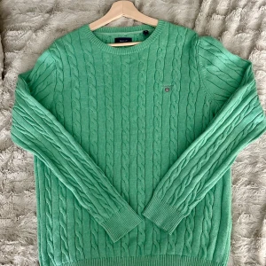 Grön kabelstickad tröja från GANT - Snygg grön kabelstickad tröja från GANT i 100% bomull. Klassisk rund halsringning, ribbade muddar och diskret GANT-logo på bröstet. Perfekt för dig som gillar en stilren och fräsch look med lite preppy känsla.