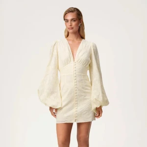 Florence Mini Dress ljusgul, Adoore - Superfin ljusgul kortklänning i storlek 34 från Adoore med puffiga långärmar, djup v-ringning och dekorativa knappar framtill. Klänningen har blommigt mönster och markerad midja för en snygg siluett. Helt slutsåld på Adoores hemsida och aldrig använd. Perfekt för sommarens alla tillfällen!
