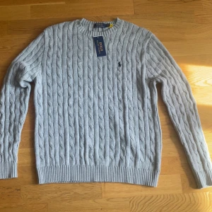 Ljusgrå stickad tröja från Polo Ralph Lauren - Säljer en ljusgrå stickad tröja från Polo Ralph Lauren med klassiskt kabelstickat mönster och broderad logga på bröstet. Jag e cirka 180 och väger 75 kg använd cirka 4-5 gånger 