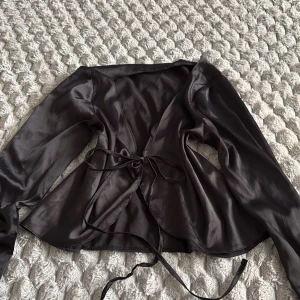 Svart omlottopp i satin från Chiquelle - Säljer en svart omlottopp från Chiquelle i glansigt satinliknande material. Toppen har långa ärmar och knytband i midjan för en snygg passform. Perfekt för dig som gillar stilrena och trendiga plagg med lite extra shine.