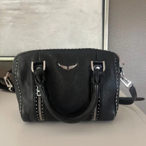 Svart Zadig & Voltaire handväska i skinn - Cool svart handväska från Zadig & Voltaire i grymt skinn med silverfärgade detaljer, nitar längs kanterna och dubbla dragkedjor framtill. Axelrem med broderad logga och snygg metallvinge på framsidan. Perfekt storlek för det viktigaste.