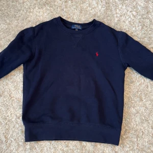 Mörkblå sweatshirt från Polo Ralph Lauren - Klassisk mörkblå sweatshirt från Polo Ralph Lauren med rund halsringning och röd broderad logga på bröstet. Tröjan har långa ärmar och ribbade muddar vid ärmslut och nederkant. Perfekt för en chill och stilren look.