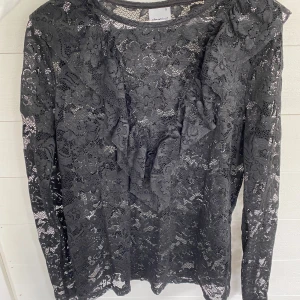 Svart spetsblus med volang från Gina Tricot - Supersnygg svart blus i spets från Gina Tricot med lång ärm och volangdetalj framtill. Blusen har ett blommigt spetsmönster och är genomskinlig, perfekt att styla med ett linne under. Ger en cool och elegant vibe till din outfit.