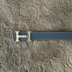 H bälte - Snyggt blått skärp från H med ikoniskt H-spänne i silverfärg. Bältet är gjort i slätt skinn med vita kontrastsömmar längs kanterna. Perfekt accessoar för att lyfta din outfit med en lyxig touch och klassisk design.