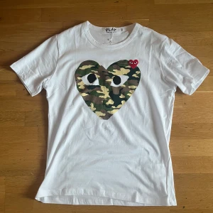 Comme des Garçons Play camo t-shirt - Vit t-shirt från Comme des Garçons Play med stort hjärta i camouflage på bröstet och ett litet rött broderat hjärta med ögon. Klassisk rund hals och korta ärmar. Skön passform i mjuk bomull, perfekt för en cool streetstyle-look.