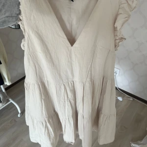 Beige volangklänning från H&M - Supersöt kort beige klänning från H&M med volangdetaljer på axlarna och djup v-ringning. Klänningen har en luftig passform och är sydd i ett lätt bomullstyg, perfekt för varma dagar. Den har flera lager och en romantisk vibe.