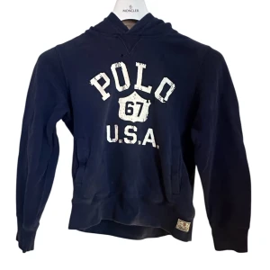 Mörkblå hoodie Polo Ralph Lauren - Mörkblå hoodie från Polo Ralph Lauren med vit tryckt text 'POLO 67 U.S.A.' på bröstet. Storlek M 10-12 år, två små hål men annars bra skick! Klassisk känguruficka och huva med amerikanskt flaggmönster på insidan. Skön passform och perfekt för chill dagar.