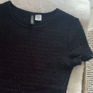 Svart smockad topp från H&M Divided - En svart smockad topp från H&M Divided med rund halsringning och korta ärmar. Tyget är elastiskt och har en strukturerad, ribbad känsla som ger en snygg passform. Perfekt att styla med jeans eller kjol. 