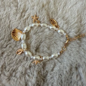 Pärlarmband med snäck- och stjärnberlocker - Superfint armband med oregelbundna vita pärlor och detaljer i guld. Armbandet har berlocker i form av snäcka, sjöstjärna och små ovala former, vilket ger en somrig och trendig vibe. Justerbar kedja i guld för perfekt passform.