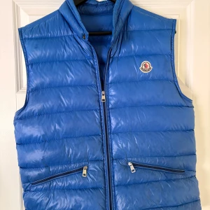Moncler väst - Snygg blå dunväst från Moncler med stilren design och hög kvalitet. Västen har två dragkedjefickor framtill och klassisk Moncler-logga på bröstet. Perfekt att slänga över en hoodie för en cool look. Klockrent för sommaren och för dig som vill sticka ut lite. Kan gå ner i pris lite vid snabb och smidig affär.