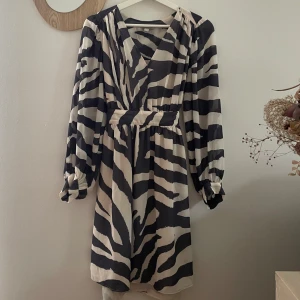 Zebramönstrad kortklänning med knyt - Snygg kortklänning med svartvitt zebramönster och v-ringning. Klänningen har långa ärmar med lätt puff och ett brett knytband i midjan som ger en markerad siluett. Perfekt för dig som vill sticka ut med en trendig look. Passar perfekt till ett par boots
