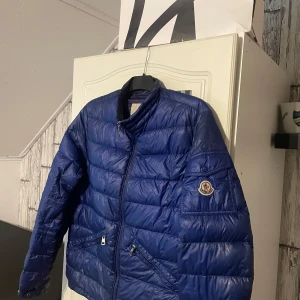 Blå Moncler sommarjacka herr - Säljer en blå dunjacka från Moncler med klassisk logga på ärmen. Jackan har quiltad design, dragkedja framtill och två fickor med dragkedja. Perfekt för kalla dagar och riktigt snygg till streetwear-stilen. Finns inget kvitto men fås med i NK påse. Jackan har använts men är fortfarande i bra skick, eftersom den ändå är använd och inte finns något äkthetsbevis säljs den lite billigare