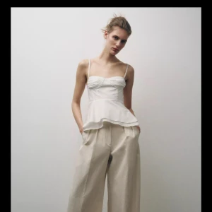 Vit peplum linnetopp med knytband - Supersöt vit linnetopp med smala axelband och peplumvolang. Toppen har rynkade kupor och knytband i ryggen för extra detalj. Perfekt för en fräsch och somrig look, lätt att matcha med både jeans och kjol.