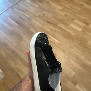 Svarta sneakers med glitter från Liu Jo - Snygga svarta sneakers från Liu Jo med glittrig tå och vita sulor. Skorna har svarta snören och en metallogo på sidan. Ovandelen är i skinnliknande material och glitterdetaljerna ger en extra touch. Perfekta för dig som vill sticka ut lite extra.