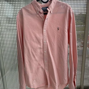 Rosa skjorta från Polo Ralph Lauren M - Snygg rosa skjorta från Polo Ralph Lauren i slim fit med klassisk button down-krage och broderad logga på bröstet. Skjortan är långärmad och har vita knappar framtill. Perfekt för dig som gillar stilrena och tidlösa plagg.