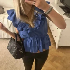 Blå volangblus från H&M - Supersöt blå blus från H&M med volanger längs axlar och ringning. Modellen är ärmlös och har resår i midjan för en snygg passform. Perfekt att styla med jeans eller kjol för en trendig look. Storlek 36 så passar xs-s