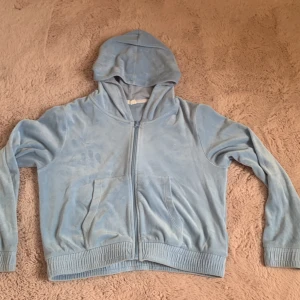 Ljusblå hoodiejacka från Lindex - Mysig ljusblå hoodiejacka från Lindex med dragkedja framtill och två fickor. Jackan är i mjuk velour med ribbade muddar och huva, perfekt för chill dagar eller när du vill vara extra comfy.
