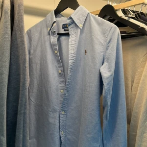  Ralph Lauren - Klassisk ljusblå skjorta från Ralph Lauren med broderad logga på bröstet. Skjortan har lång ärm, knappar hela vägen och button-down krage. Custom fit-modell i mjuk bomull, perfekt till många olika tillfällen. Skick 9-10 mitt pris 499