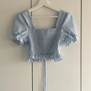 Ljusblå blommig croppad blus från SHEIN - Supersöt ljusblå croppad blus från SHEIN med små vita blommor, puffärmar och volangdetalj nertill. Smockad över bysten och knytband bak. Perfekt för vår och sommar! Inte använd.