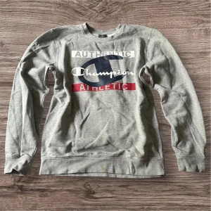 Grå sweatshirt från Champion - Säljer en grå sweatshirt från Champion med tryck i vitt, blått och rött framtill. Tröjan har lång ärm och klassisk passform. Perfekt för en avslappnad stil och enkel att matcha med jeans eller joggers.