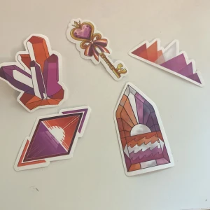 Färgglada klistermärken med kristall- och solnedgångsmotiv - Säljer ett set med fem unika klistermärken i lila, orange och rosa toner. Motiven inkluderar kristaller, en nyckel med hjärta, stiliserade solnedgångar och geometriska former. Perfekta för att dekorera laptop, mobil eller anteckningsbok.
