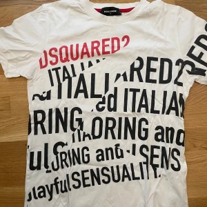 Vit t-shirt med tryck från Dsquared2 - Säljer en vit t-shirt från Dsquared2 med stor textprint i svart och rött över hela framsidan. T-shirten har rund halsringning och är tillverkad i 100% bomull. Perfekt för dig som gillar statement-plagg och streetstyle.