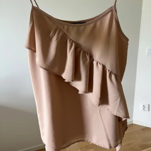Beige linne med volang - Säljer ett beige/ljusrosa linne med tunna axelband och snygg volangdetalj framtill. Perfekt för varma dagar eller att styla lager på lager. Luftigt och lätt material som känns skönt mot huden.