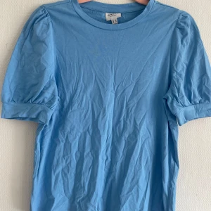 Blå topp med puffärm från Vero Moda - Säljer en ljusblå topp från Vero Moda med puffärmar och rund halsringning. T-shirten har en enkel och stilren design med korta ärmar som har lätt volym. Perfekt att styla till jeans eller kjol! ( Stryks innan postningen )