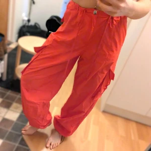 Röda cargopants med dragsko - Säljer ett par röda cargopants med flera fickor på sidorna och dragsko vid benslut. Byxorna har normal passform och stängs med knapp och dragkedja. Perfekta för en avslappnad och trendig stil.