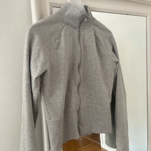 Grå ziptröja med hög krage  - En stilren grå ziptröja med hög krage och långa ärmar. Tröjan har ribbade muddar och midja samt dragkedja framtill. Perfekt för lager-på-lager och enkel att matcha med det mesta. Populär tröja från Nelly. Lite mindre i storlek så passar en M. Endast testad/använd några timmar. 