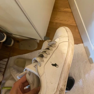 Vita sneakers från Polo Ralph Lauren - Säljer ett par klassiska vita sneakers från Polo Ralph Lauren med svart logga på sidan och Polo-tryck på hälen. Skorna har snörning och rund tå, perfekt till vardagsstilen. Riktigt sköna lite smutsiga men går o få bort med lite vatten, köpta för 1,4 på sportslab 