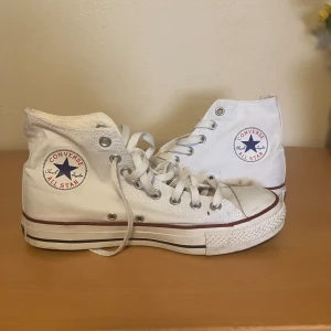 Vita Converse sneakers - Klassiska vita Converse All Star high-top sneakers med snörning och rund tå. Skorna har den ikoniska loggan på sidan och en platt sula med röd rand runt om. Perfekta för en avslappnad och tidlös stil.