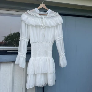 Vit volangklänning med spetsdetaljer - Romantisk vit klänning med volanger och spetsdetaljer. Klänningen har långa ärmar med volang och smock i midjan för en feminin siluett. Kjolen är kort med dubbla volanglager och omlottdetalj vid halsen. Klänningen har en liten fläck på ett av snörena, men det är inget som syns så tydligt eller som man tänker på. Har köpt den på plick och använt den som en del av en piratkostym till halloween. Säljer även korsetten jag använde till i en annan annons. 