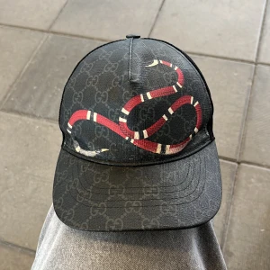 Svart keps från Gucci med orm-motiv - Snygg svart keps från Gucci med diskret GG-mönster och en stor röd och vit orm på framsidan. Kepsen har mesh på sidorna för extra ventilation och justerbar passform. Perfekt accessoar för att sticka ut i mängden.