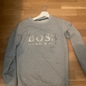 Grå sweatshirt från Hugo Boss - Säljer en stilren grå sweatshirt från Hugo Boss med logga i vitt framtill. Tröjan har rund halsringning och långa ärmar. Sällsynt tröja som är svår att få tag på! 