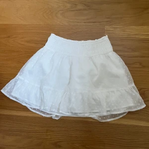 Vit kjol med volang från Abercrombie Kids - Supersöt vit kjol från Abercrombie Kids med volangkant och diskret prickigt mönster. Kjolen har även inbyggda shorts!