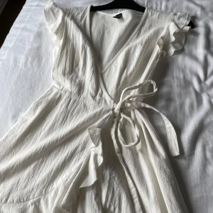 Vit omlottklänning med volangdetaljer - Säljer en söt vit omlottklänning med knyt i midjan och volangdetaljer längs ärmar och kjol. Klänningen har korta ärmar och en v-ringad halsringning. Perfekt för sommarens alla tillfällen eller kanske en konfirmation!! 