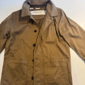 Brun overshirt från Selected Homme - Säljer en brun overshirt från Selected Homme med klassisk krage och tre stora fickor framtill. Jackan har knäppning med svarta knappar och är tillverkad i ett slitstarkt bomullstyg. Perfekt för lager-på-lager-stil.