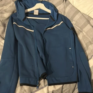 Blå Nike hoodiejacka med dragkedja - Säljer en blå hoodiejacka från Nike med hel dragkedja och huva. Jackan har vita detaljer och Nike-logga på bröstet. Tröjan sitter som en xxs