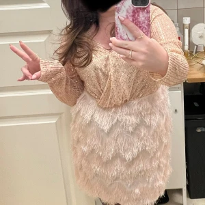 Glittrig champagnefärgad klänning med fransar - Säljer en glittrig kortklänning i champagnefärg med långa ärmar och v-ringning. Överdelen är täckt av paljetter och kjolen har flera lager av fransar som ger en festlig känsla. Använd endast en gång. 