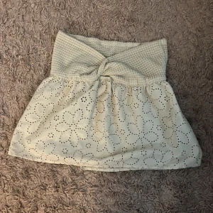 Vit kortkjol med broderade blommor och virkad midja - Supersöt vit kortkjol med broderade blommor och hålmönster. Kjolen har en bred, virkad midja med twistdetalj framtill som ger en unik look. Perfekt att styla med en enkel topp för en fräsch sommarstil.