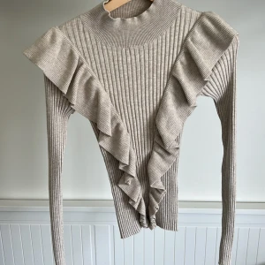 Beige ribbad tröja med volang från Lindex - Säljer en beige ribbad tröja från Lindex med långa ärmar och snygga volangdetaljer framtill. Tröjan har hög hals och är perfekt för dig som gillar en trendig och stilren look. Passar till många olika outfits! Priset går att diskutera 