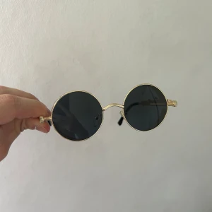 Runda solglasögon med guldfärgade bågar - Snygga runda solglasögon med tunna guldfärgade metallbågar och mörka svarta glas. Skalmarna är svarta och ger en cool retrostil. Perfekta för att lyfta din look med en unik touch.