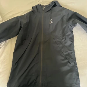 Svart regnjacka från Haglöfs - Säljer en svart regnjacka från Haglöfs med huva och hel dragkedja framtill. Jackan har två snedställda fickor och minimalistisk design, perfekt för blöta dagar. Märke och storlek syns tydligt på insidan.
