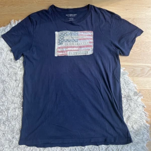 Mörkblå t-shirt med flaggtryck från Ralph Lauren - Säljer en mörkblå t-shirt från Ralph Lauren med ett stort amerikanskt flaggtryck och broderade detaljer på bröstet. T-shirten har rund hals och korta ärmar. Perfekt för en avslappnad stil. Notera att jag säljer 2 sånnahär tshirts på min profil. Hör gärna av dig om du har frågor!