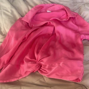 Rosa knytblus från Lindex Kids - Säljer en snygg rosa blus från Lindex Kids i satinliknande material. Blusen har korta ärmar, klassisk krage, knappar framtill och en knytning nedtill för en trendig look. Perfekt för dig som vill sticka ut lite extra!