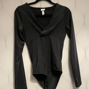 Svart långärmad bodysuit från H&M - Säljer en svart långärmad bodysuit från H&M med djup v-ringning och åtsittande passform. Perfekt att styla med jeans eller kjol för en trendig look. Materialet är stretchigt och ger en smickrande siluett.