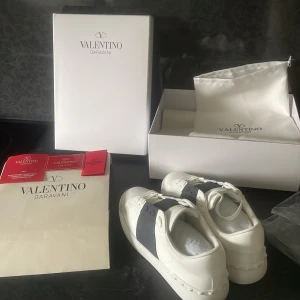 Valentino Garavani Open sneakers med blå detalj - Säljer ett par vita Valentino Garavani Open sneakers med bred svart rem över mitten. Skorna har rund tå, platt sula och klassisk snörning. Kommer med originalkartong, dustbag och påse. Perfekta för dig som gillar lyxiga och stilrena sneakers.