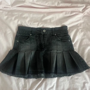 Svart plisserad jeanskjol - Säljer en svart kort jeanskjol med plisserad design och rå kant nertill. Kjolen har klassiska jeansfickor fram och bak, bälteshällor och stängs med knapp och dragkedja. Perfekt för dig som gillar en trendig stil. 